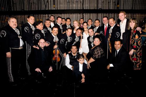 A group of over 20 people, many in black mariachi uniforms and other formal dress, some holding violins, pose, smiling.