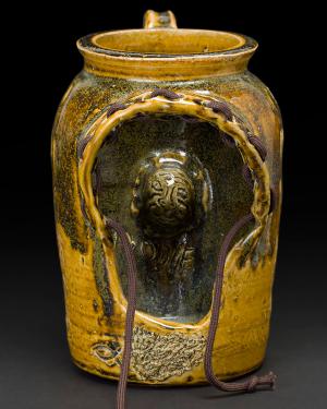 Yellow glazed ceramic jug with a cutout in the side, resembling the hood of a sweatshirt. Inside, carved in the inner wall of the jug, is the face of a boy. Brown cloth pullstrings come out of the hoodie shape.