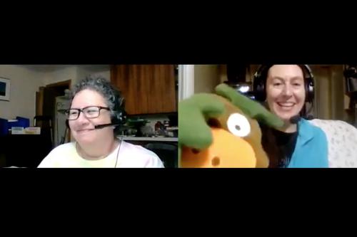 A screenshot of a two-person Zoom call. On the right, a woman wearing glasses and a headset smiles, on the left, a woman wearing a headset holds a big puppet up to the screen while laughing.