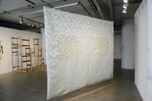A light-colored embroidered sheet hangs from the ceiling in an art gallery.
