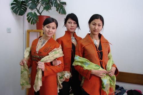 Three women look at the camera and smile in orange felt outerwear. There are light green scarves on their arms.