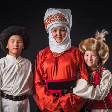 An adult and two young children are dressed in traditional garments from Kyrgyzstan. They smile at the camera against a black background.