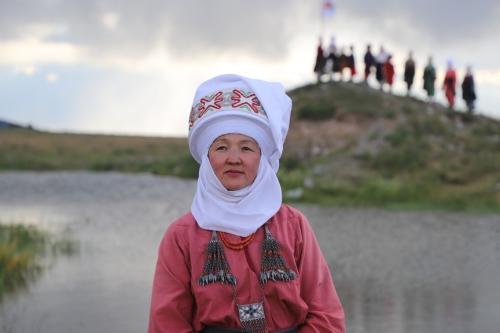 An adult who is dressed in traditional garments from Kyrgyzstan stands outside and looks past the camera. A group of adults stand on a hill in the background.