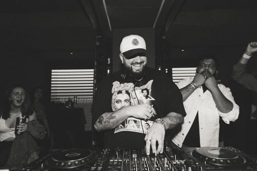An Arab man with dark beard and tatooed arms smiles, standing at a DJ mixer. People around him whistle and cheer.