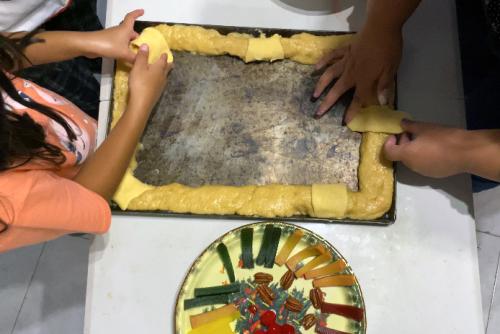 From above, two people place rectangles of pale dough on top of a rectangular ring of light brown dough, arranged on a rectangular baking pan. The toppings of fruit and nuts are on a plate nearby.