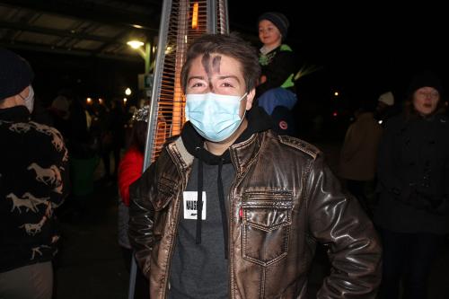 A person in a leather jacket and surgical mask, with a smudge of ash on their forehead.