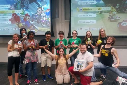 Eleven people pose in front of two projector screens showing video game option menus. Each person holds a prize, mostly small plush toys.