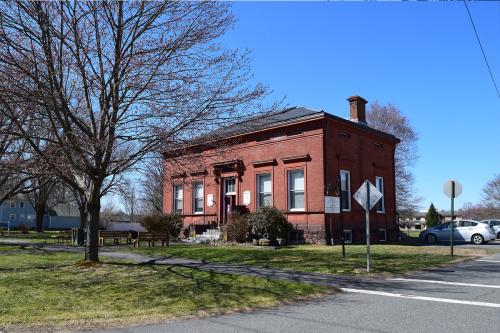 Same red brick building, with different trees that are also bare. A street has been paved with street signs and cars parked nearby. Color photo.