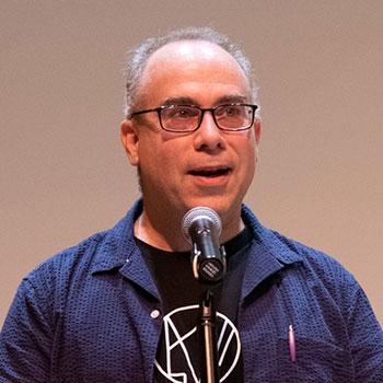 Dovid Braun speaking into a microphone, wearing blue shirt and glasses.
