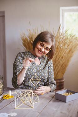 A woman smiles, seated at a work table and holding up a small octahedron made of straw. More shapes sit in front of her, and a bouquet of cut, dried straw behind her.