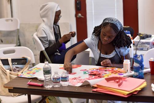 A young woman works with colorful pieces of paper over a wooden table inside of a studio space. A young man wearing a white hoodie sits behind her working with his hands.