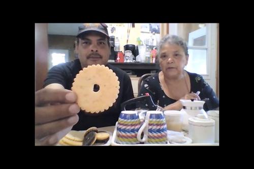 A man wearing a black t shirt and baseball cap holds a circular cookie up to the camera. An older woman with short grey hair sits next to him.