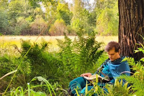 A person wearing a bright turquoise robe reads at the base of a tree, surrounded by ferns, with a field and forest in the background.