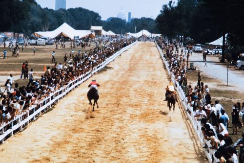 Two horse jockeys race down a dirt track on the National Mall, with crowds gathered behind white fences on either side. In the distance is the U.S. Capitol Building.