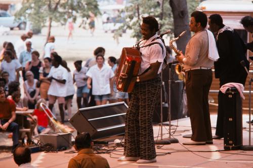 A man performs on an outdoor stage for a crowd of dancing people, playing the piano accordion and accompanied by his band.