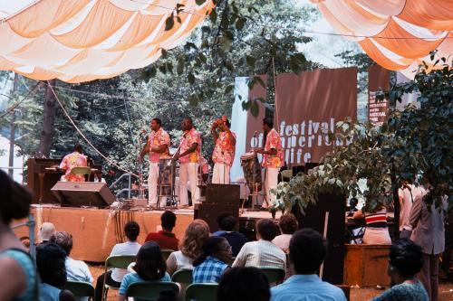 A band in matching pinkish floral shirts and white pants on an outdoor stage, under a matching pink tent. A crowd sits watching them.