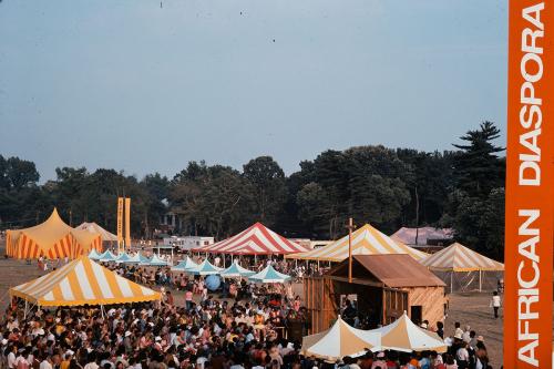 Wide shot of colorful striped festival tents and crowds of people on an open lawn. To one side, a tall, vertical, orange sign reading AFRICAN DIASPORA.