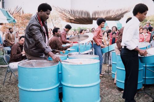 Outdoors, several Black men play steel drums painted bright blue for a crowd.