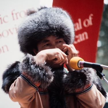 A person in fur lined coat and hat performs on an outdoor stage.