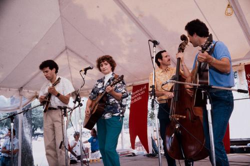 Four people on stage, three men and a woman, with a banner behind them for the Festival of American Folklife. The woman plays guitar; the men play mandolin, upright bass, and guitar.