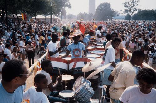 Percussionists on a truck bed, moving through a crowd of people. The base of the Washington Monument is in the background.