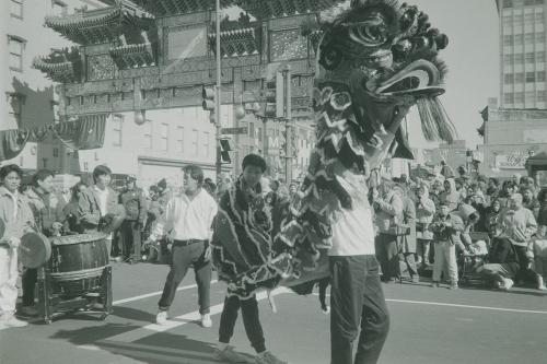 Two young men enliven a Chinese dragon costume, the the one in front raising its head high. Behind them, other young men play percussion instruments, and a crowd forms under the Chinese archway over the street.