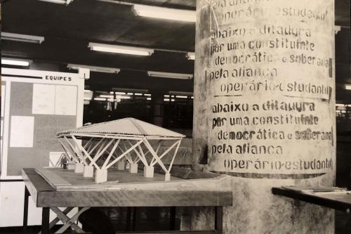 A poster in Portuguese on a column, beside an architectural model on a table. Black-and-white photo.