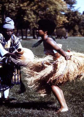 A person dances outdoors, wearing a thick grass skirt and cloth bandeau top. Another person plays a gourd percussion instrument.