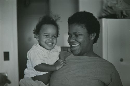 A woman with short Afro smiles looking at the baby she holds up with one arm. The baby smiles widely at the camera. Black-and-white photo.