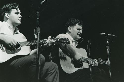 Two men, the one of the left younger, sit and play acoustic guitar on stage.