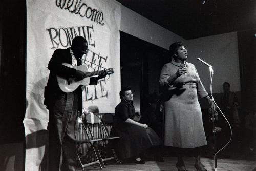 A man on an acoustic guitar and a woman singer perform on a low stage. A hand-painted banner behind them reads Welcome Ronnie, Lee, Pete. Black-and-white photograph.
