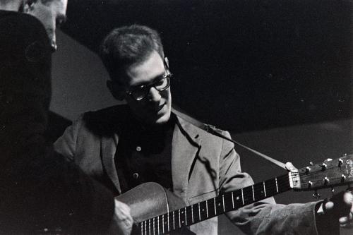 A young man wearing glasses and a blazer tunes up an acoustic guitar. Black-and-white photograph.