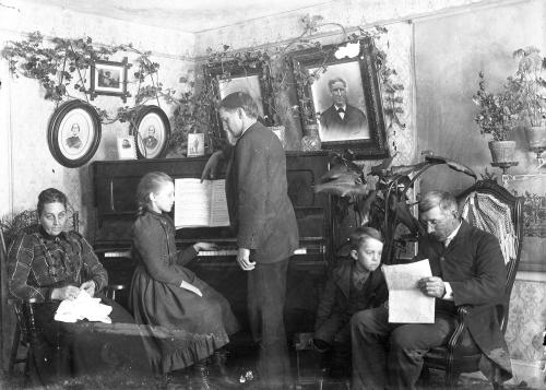 A family of five in a parlor. Daughter plays on piano, while the father (presumably) points at her sheet music. The mother has a textile in her lap. Grandfather and grandson read a paper. Old black-and-white photo.