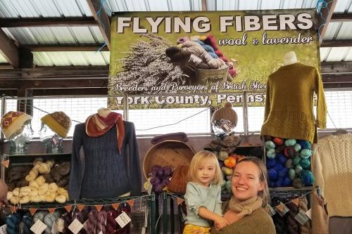Under a banner for Flying Fibers, a display of dyed yarn and sweaters and a woman holding a toddler.