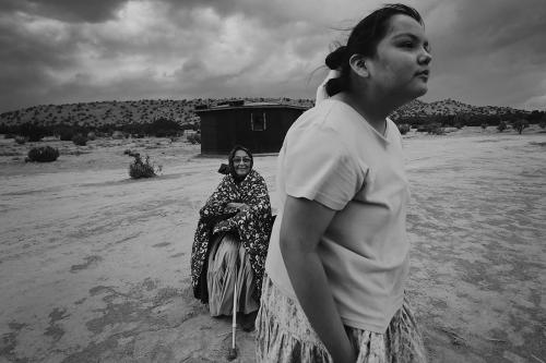 A young girl gazes into the distance in the foreground, with an elder woman seated behind her, smiling slightly, in a desert landscape. Black-and-white photo.