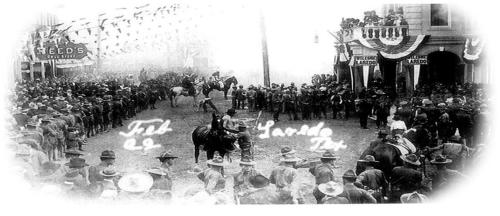 Old black-and-white image of people on horseback surrounded by crowds of people on a city street. Handwritten script in white reads Laredo Tex.