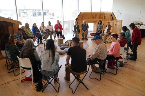 Twenty people seated in a circle in a museum gallery space, lined on one side with bright windows. In the center of their circle are rocks and pieces of wood, arranged like a fire pit.