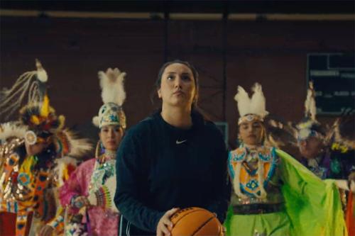 A woman in black athletic clothes readies herself to shoot a basketball, while others dressed in elaborate regalia dance behind her.