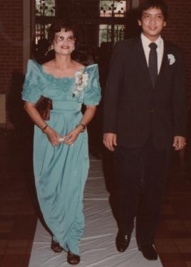A Filipino man and woman walk down a white carpet, wearing a dark suit and a turquoise dress with pointed sleeves, each with a bluish-white corsage.