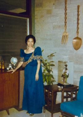 Inside a home, a woman with short dark hair poses wearing the blue dress with gold embroidery and pointed sleeves. On the wall above her hang a set of giant, decorative fork and spoon.