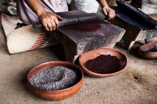 A pair of hands rolling a pin on a stone to create a purple paste. Beside the stone is a basket full of blueish dried bugs.