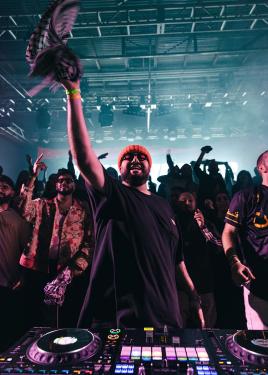 A man at a DJ mixer, with a crowd waving behind him, raises one arm to wave a keffiyeh scarf.