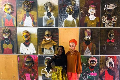 Two women pose in front of a wall of abstract portraits arranged in a grid.