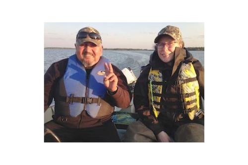 A man and a woman sit next to each on a boat, other smiling. They both are wearing baseball caps and lifejackets and in the background is open water.