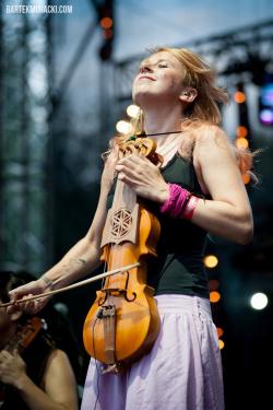 A woman stands on stage, head tilted up with eyes closed. She holds a violin-like instrument straight up by the neck in front of her torso, playing it with a bow with the other hand.