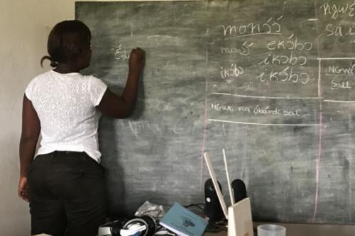 A woman stands at a chalkboard writing in Iyasa. Her hair is pulled back into a bun, and she wears a white shirt and black pants.