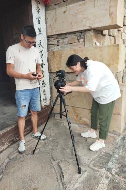 Two people stand together preparing an audio and video recording. A man holds a small audio recorder and a woman stands, bent over, adjusting a tripod.