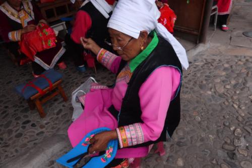Balanced on a stool low to the ground sits a woman in the midst of an embroidery project. She wears a traditional Qiang dress including a white head wrap, pink tunic, and black vest.