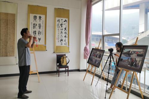 A man stands in the midst of performing a short tune on a Qiang flute. Across from him, a young woman stands with a camera on a tripod, recording him.