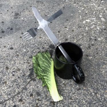 Closeup of a cross made from a metal fork and knife taped together, held up in a mug, sitting next to a lettuce leaf on a sidewalk.